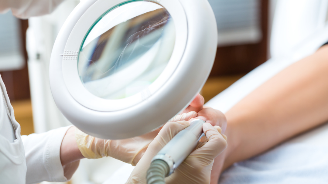 Image is a close up of a salon manicurists hands in gloves looking through a large round magnifying glass at a women's hand and fingers while providing a professional nail treatment